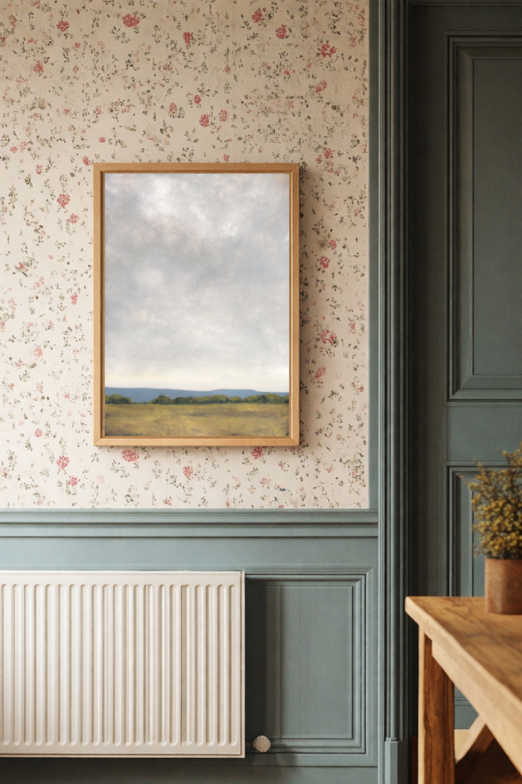 Large landscape print on floral wallpaper with a radiator and wooden table in the background.